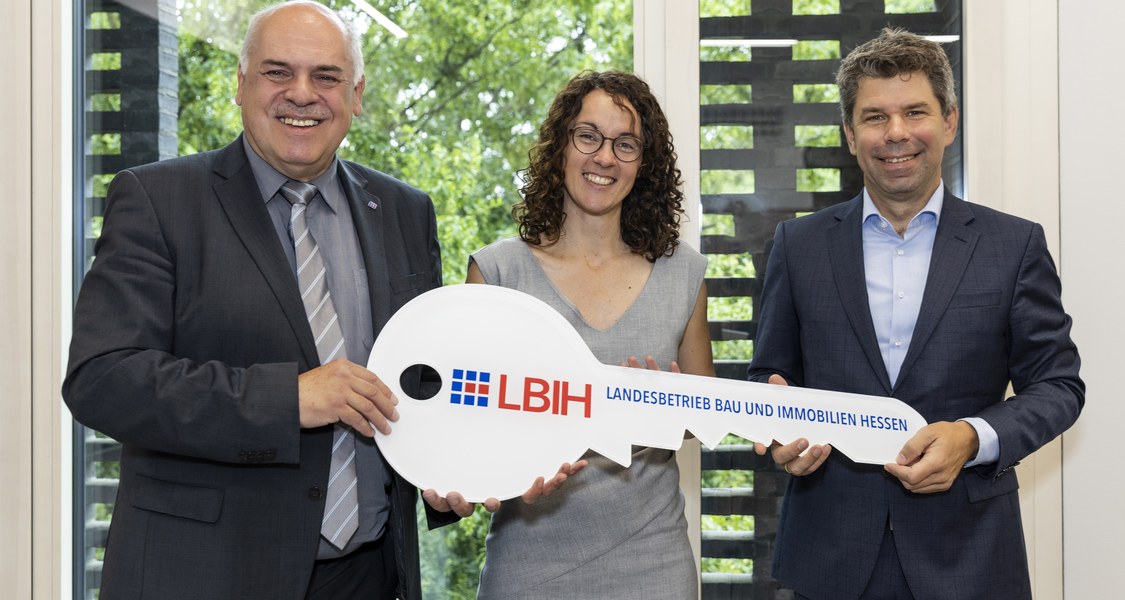 Drei Personen im Gruppenfoto vor einem Fenster mit Blick in den Botanischen Garten - ein großer symbolischer Schlüssel mit der Aufschrift Landesbetrieb Bau und Immobilien Hessen wird von allen drei Personen gehalten