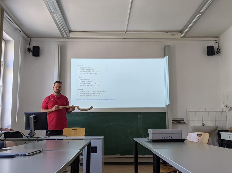 Ein Mann, der ein bronzenes, krummsäbelförmiges Schwert hält, steht vor einer Powerpoint-Präsentation über die chemischen Eigenschaften von Bronze