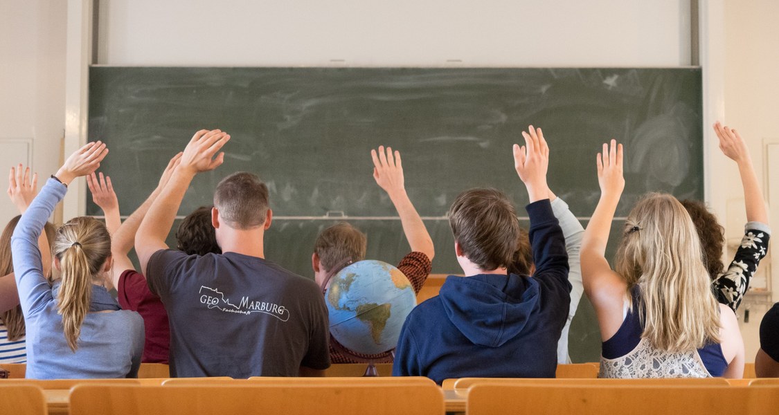 Studenten melden sich in der Vorlesung. Sie werden von hinten gezeigt. Im Hintergrund ist eine Tafel.