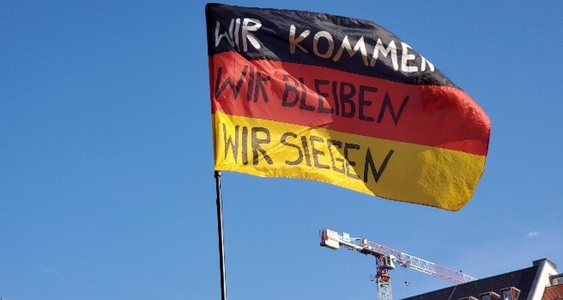 Man sieht eine Deutschlandflagge an einem Fahnenmast. Auf die schwarz-weiß-roten Streifen der Flagge wurde gut sichtbar in Großbuchstaben "Wir kommen Wir bleiben Wir siegen" geschrieben.