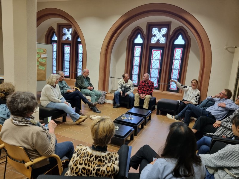 Gruppe von Menschen die auf Sesseln in einem Kreis sitzt. In der Mitte stehen kleine Tische auf denen sich Glasglocken befinden.