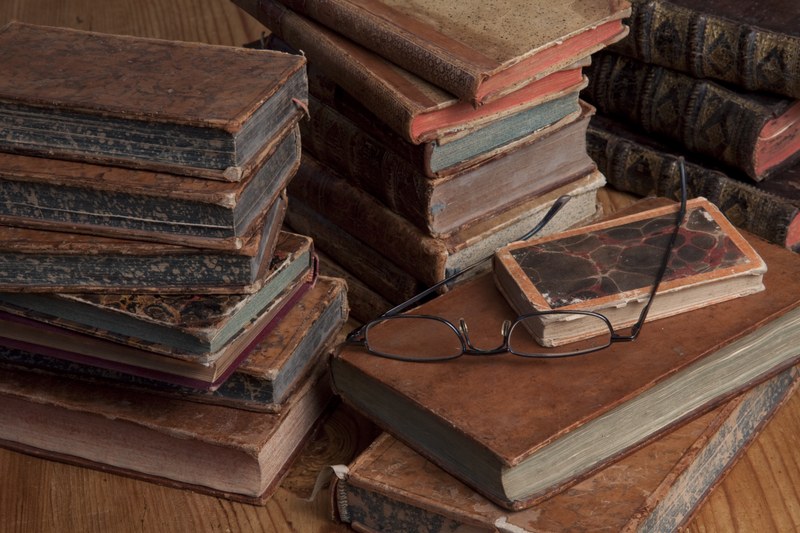 Ältere, verstaubte Bücher liegen in Stapel auf einem Holztisch mit einer Brille dazwischen.