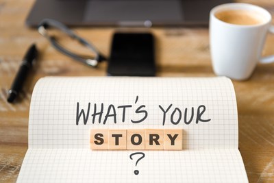 Notizbuch mit der Frage "What's your story" auf einem Schreibtisch mit Kaffeetasse, Handy, Brille und Stift im Hintergrund.