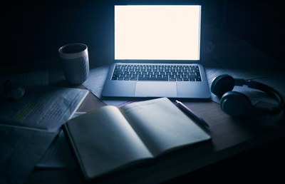 Auf einem Schreibtisch befinden sich ein Laptop mit leuchtendem Bildschirm, eine Tasse, ein Buch mit Stift und Kopfhörer.