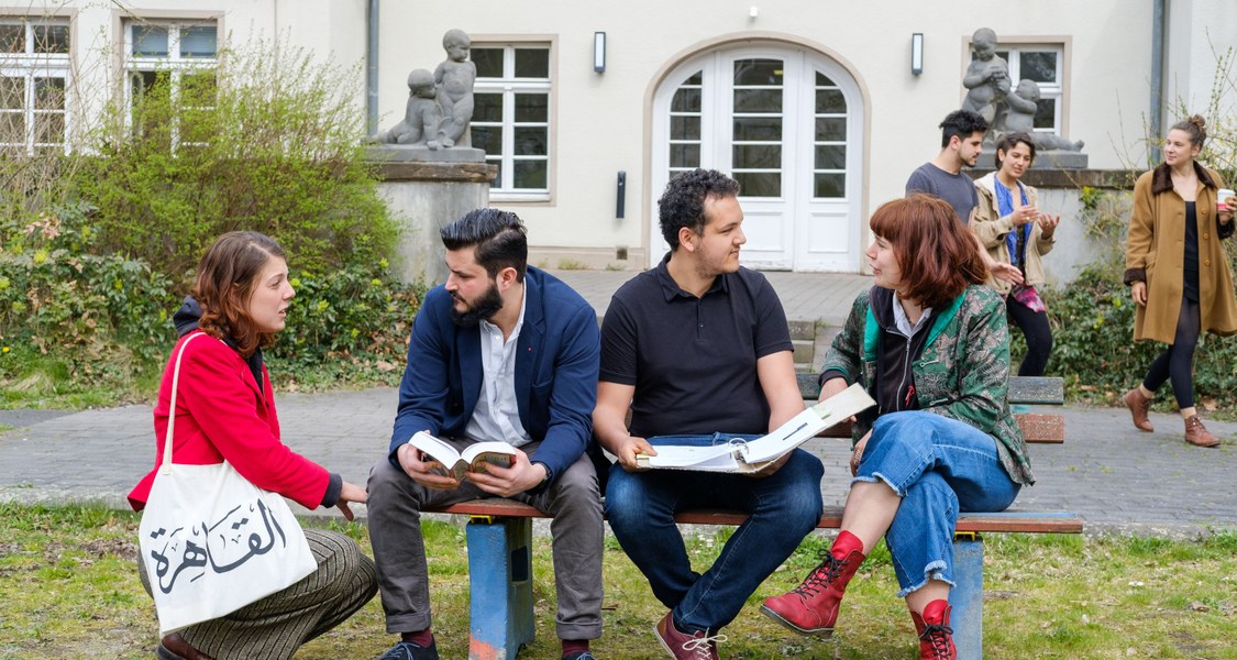 Im Hintergrund des Fotos ist ein weißes Haus zu sehen, im Vordergrund sitzen 3 Menschen auf einer Bank auf einer Wiese und unterhalten sich. Eine Frau mit rotem Pullover kniet neben der Bank und ist ebenfalls ins Gespräch einbezogen.