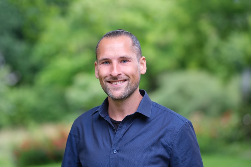 Talentscout Jonathan in dunkelblaumen Hemd vor grünem Hintergrund im Alten Botanischen Garten.