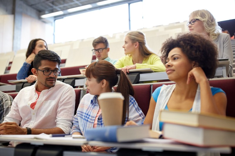 Studierende verschiedenen Ethnien sitzen in einem Hörsaal. Vor ihnen auf den Tischen liegen Bücher und Schreibunterlagen. Sie unterhalten sich miteinander.