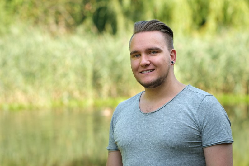 Ein junger Mann mit kurzen Haaren und Bartansatz steht in hellgrauem T-Shirt vor grünem botanischen Hintergrund und blickt lächelnd in die Kamera.