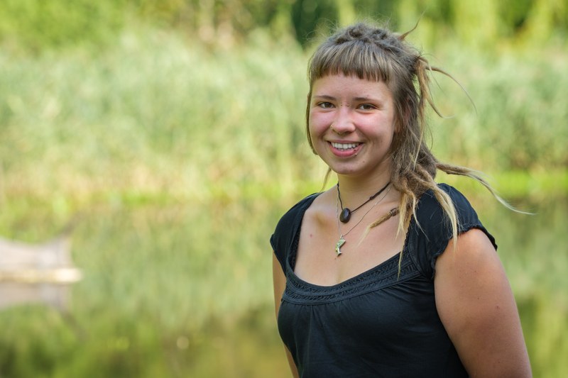 Eine junge Frau mit blonden Rastalocken, Piercings und Halskette steht im schwarzen T-Shirt vor grünem botanischen Hintergrund und blickt lächelnd in die Kamera.