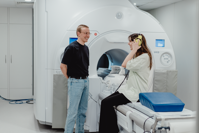 Das Foto zeigt eine Studienteilnehmerin, die vor einem MRT-Scanner (Magnetresonanztomograph) sitzt, während ein Wissenschaftler neben ihr steht.