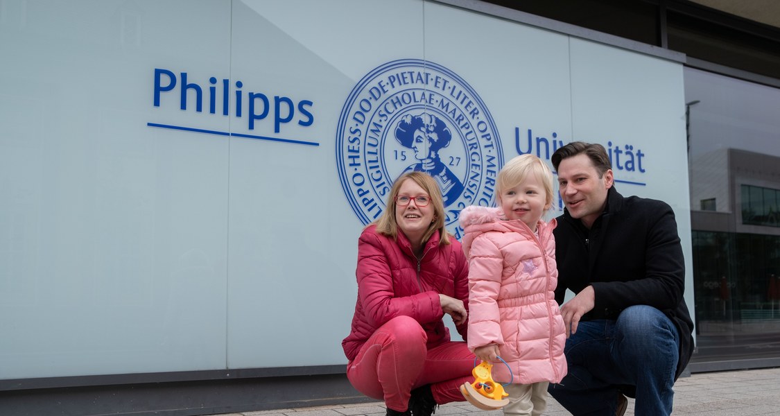 Eltern knien mit kleiner Tochter unter dem Logo der Philipps-Universität.