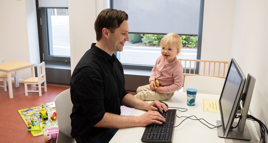 Vater arbeitet am PC, während Tochter neben ihm auf dem Tisch sitzt und lacht.