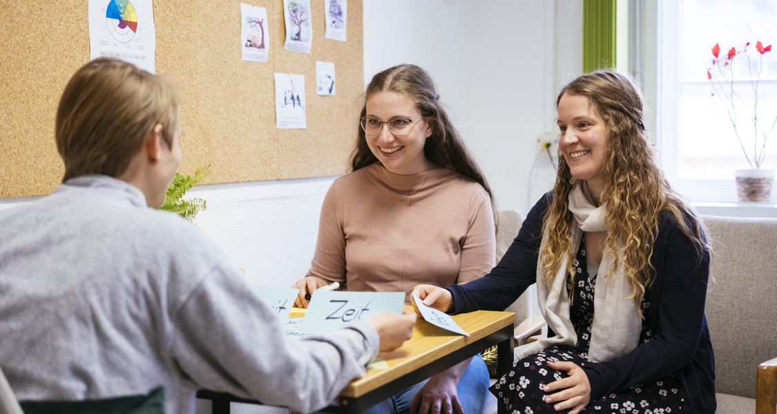 Das Foto zeigt eine Beratungssituation im Zentrum für Lehrkräftebildung. Drei Personen sitzen an einem kleinen Tisch. Zwei Frauen sitzen auf der rechten Seite des Tisches und lächeln. Sie wirken offen und aufmerksam. Ihnen gegenüber sitzt eine weitere Person mit kurzen Haaren, die dem Betrachter den Rücken zuwendet. Alle drei Personen sind in ein Gespräch vertieft.
Auf dem Tisch liegen Karten mit Namen, eine davon ist mit „Zeit“ beschriftet. Im Hintergrund ist eine Pinnwand mit mehreren ausgedruckten Blättern und Bildern zu sehen. Der Raum ist hell und freundlich gestaltet, mit einem Fenster auf der rechten Seite, durch das Tageslicht einfällt. Eine Topfpflanze steht auf dem Tisch, was zur gemütlichen Atmosphäre beiträgt