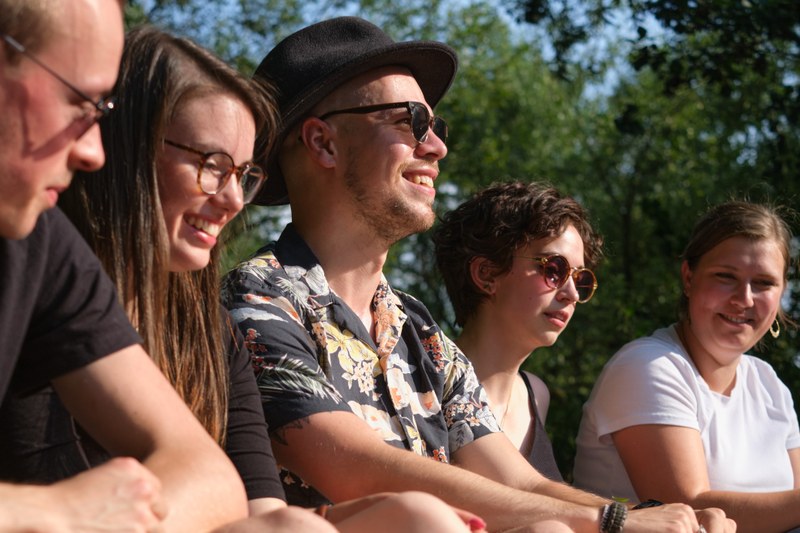 Five students are sitting barefoot by the Lahn River, smiling and chatting.