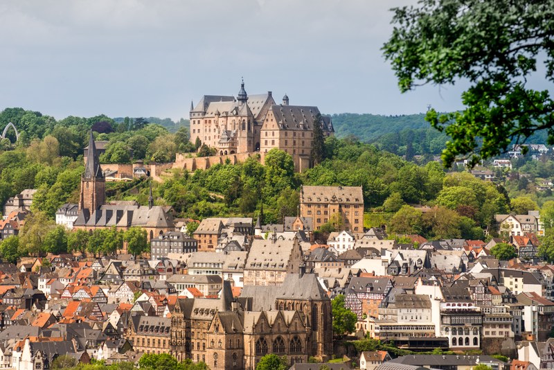 A view of the Marburg Castle, nestled above the old town and historic university buildings, surrounded by lush summer greenery.